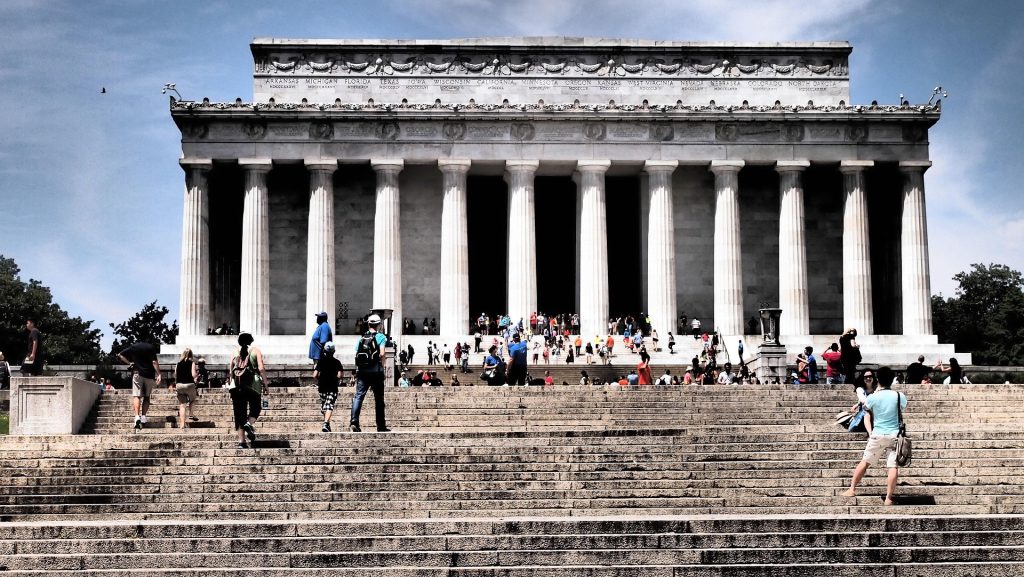 A wide view of the Lincoln Memorial in Washington D.C. under a clear blue sky, showing the massive white marble columns and the grand stone steps crowded with visitors.