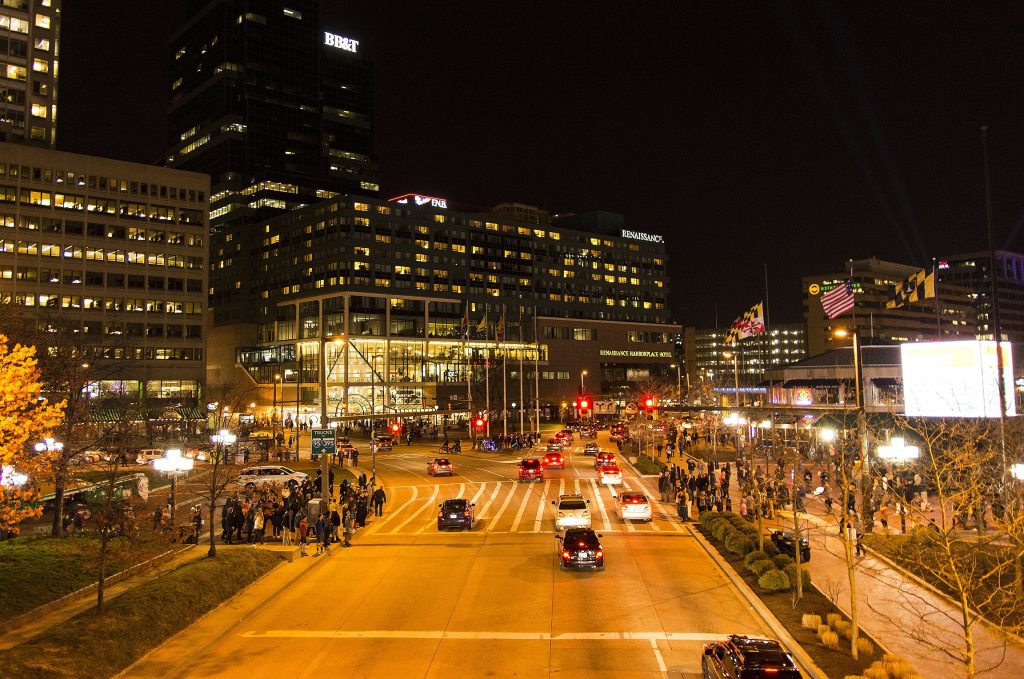 A night view of Baltimore’s Inner Harbor area with glowing streetlights, moving cars, and large buildings like the Renaissance Hotel.