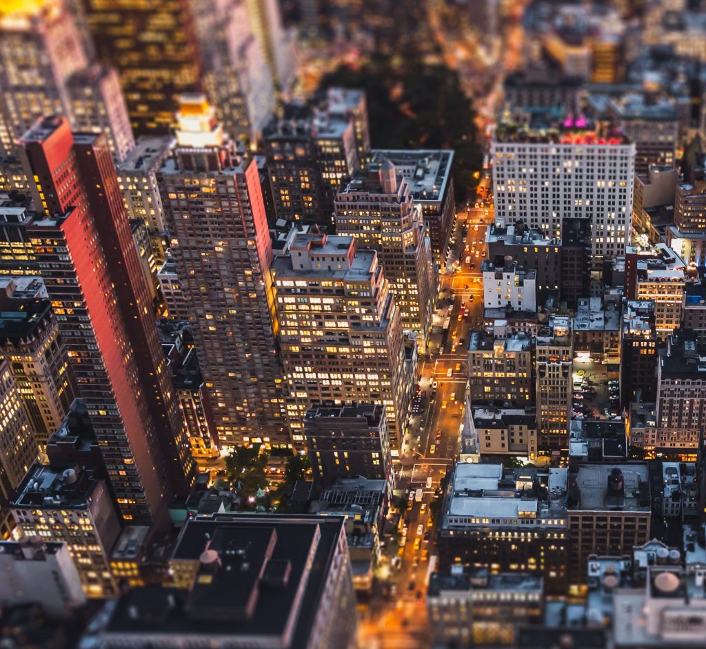 A glowing, high-angle tilt-shift view of New York City at night, showing densely packed skyscrapers and bright city lights along the streets.